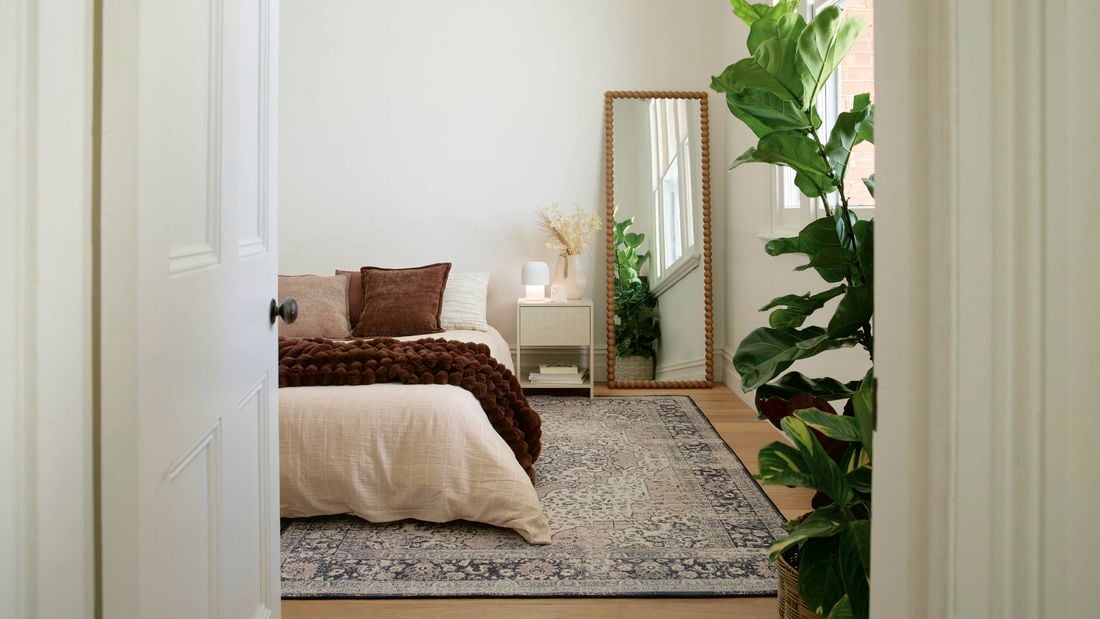 A calm bedroom viewed through a doorway, featuring a neatly made bed with neutral bedding, a patterned rug, a bedside table, a full-length mirror, and a large indoor plant by a window.