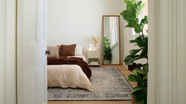 A calm bedroom viewed through a doorway, featuring a neatly made bed with neutral bedding, a patterned rug, a bedside table, a full-length mirror, and a large indoor plant by a window.