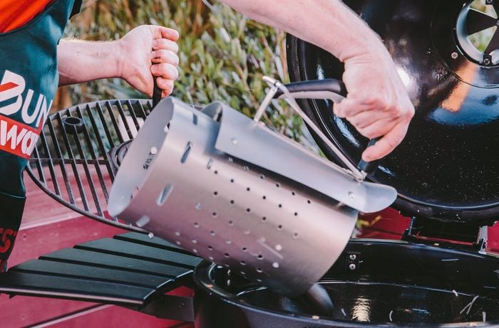 Person pouring hot charcoal briquettes into a kamado smoker