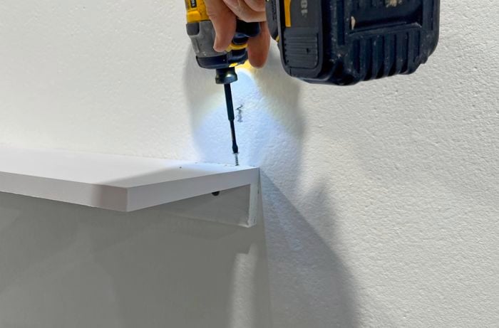 A left hand holds a white shelf against a neutral wall while the right hand uses a drill to screw the shelf into a wall batten.