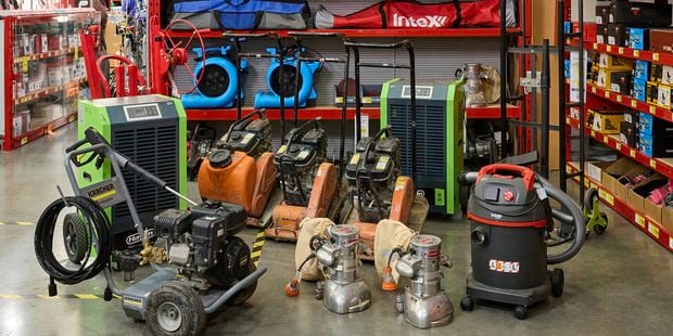 Bunnings hire section with a “Bunnings Warehouse” sign listing equipment rental prices above shelves of tools, including compactors, tile cutters, blowers, and pressure washers displayed on red racks.