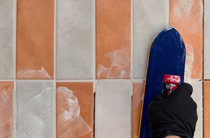 Gloved hand uses a blue grout float to spread light grey grout across orange and grey rectangular tiles.]