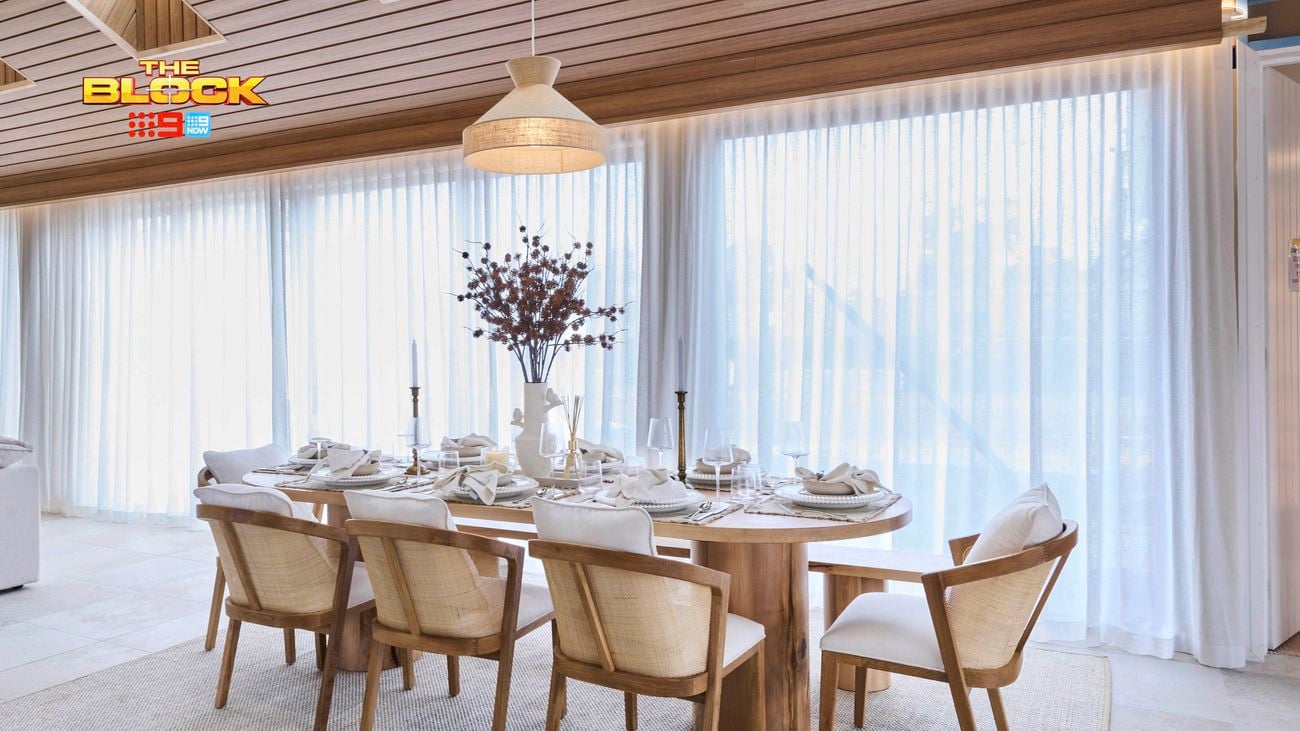 A dining room featuring a wooden table set for six, with plates, glasses, napkins, candles, and a central vase of flowers. The space is illuminated by natural light through large windows with sheer white curtains. A hanging light fixture is suspended from the wooden-paneled ceiling, and a light-colored rug lies beneath the dining set.