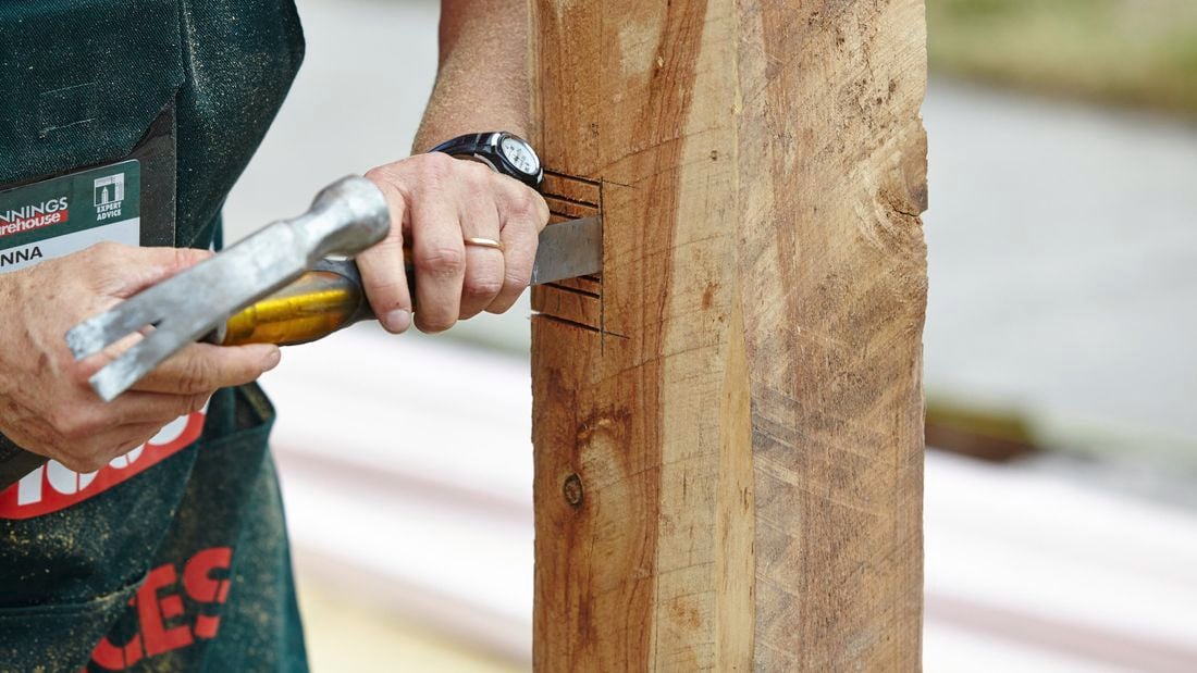 A person using a chisel and hammer to cut out slots in a fence post 