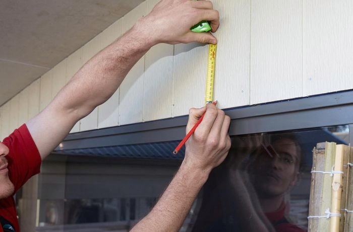 A person measuring above a window on the outside of a house