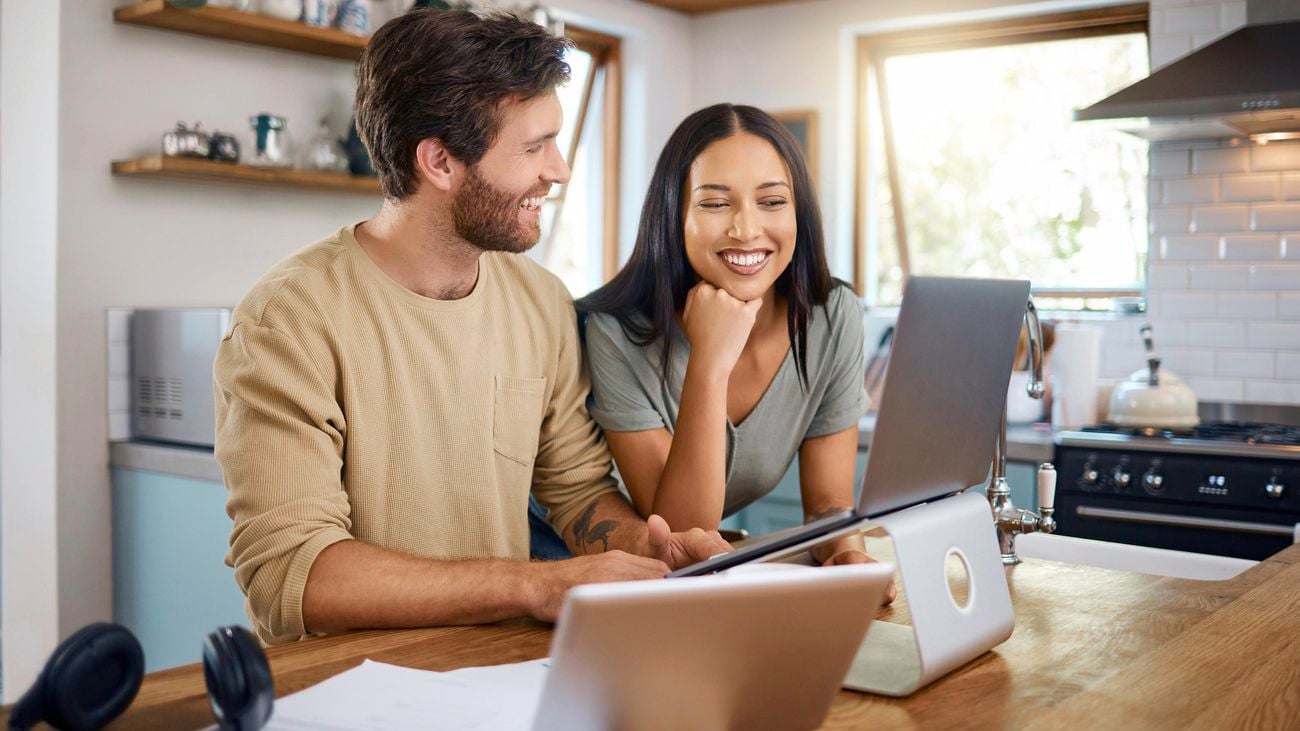 Man and woman looking at laptop and tablet on wooden kitchen bench.