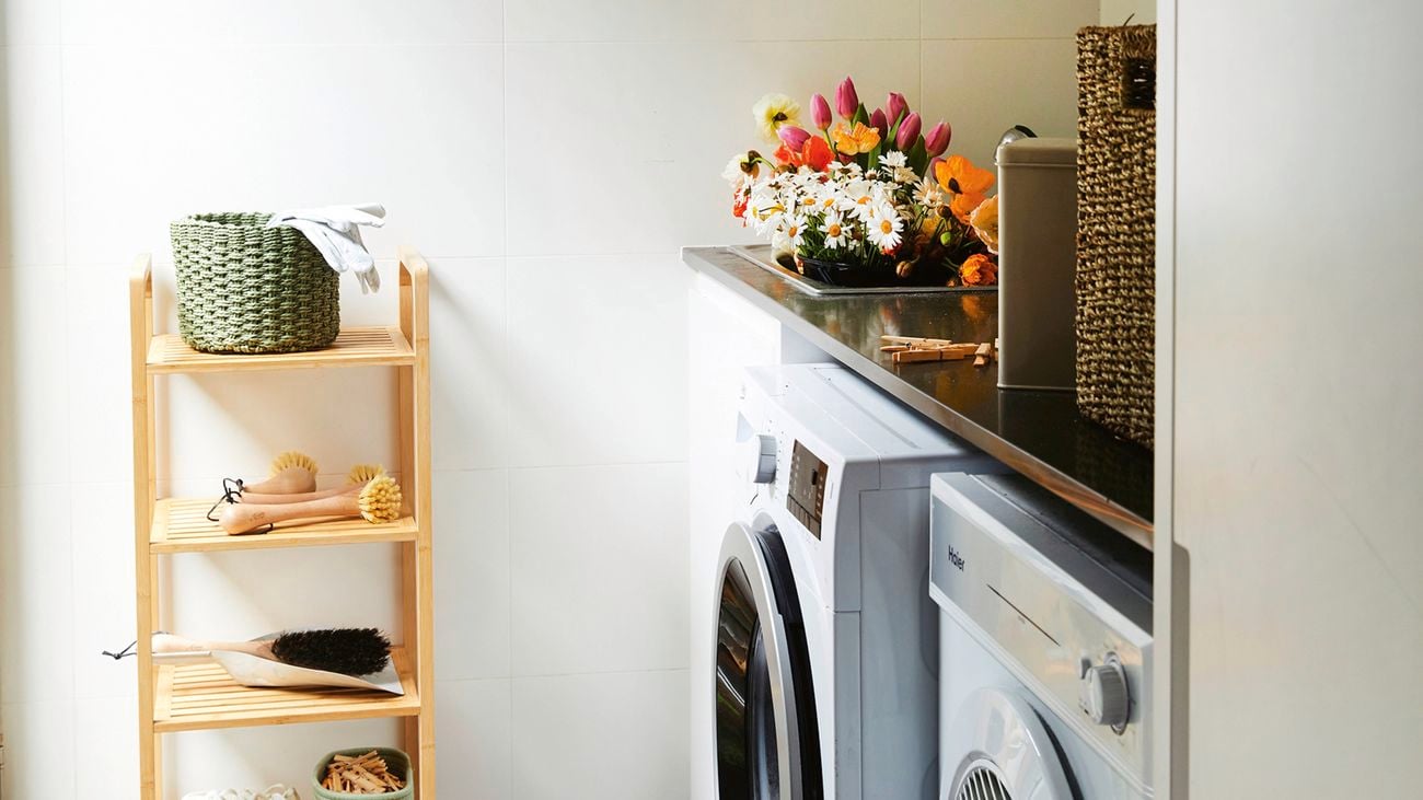 Laundry with front‑loader washer and dryer under a black benchtop. A small timber shelf holds baskets, brushes and shoes; flowers and woven baskets sit on the benchtop.