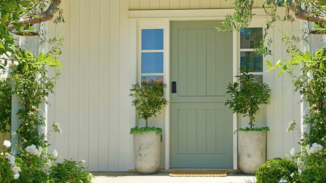 A light green front door with a black smart lock, flanked by two potted trees, and surrounded by white siding and garden foliage.