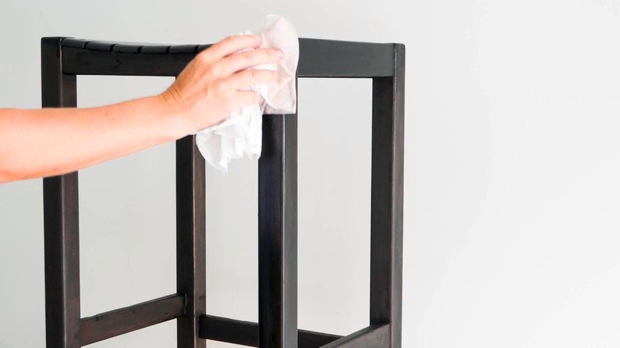 A person applying clear varnish with a cloth to a black stool from the tin on floor. 