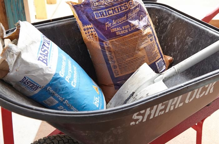 A wheelbarrow holding a shovel, a bag of concrete and a bag of bricklayer's sand