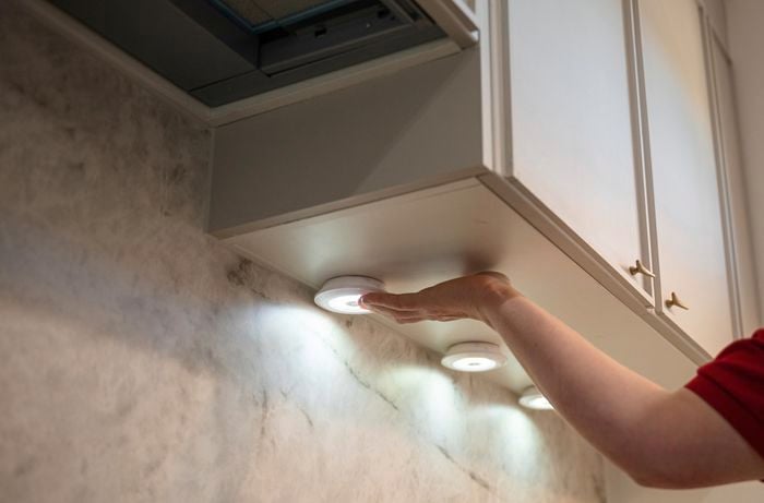 A person's hand reaching out to touch undermount lights installed beneath a kitchen cabinet.