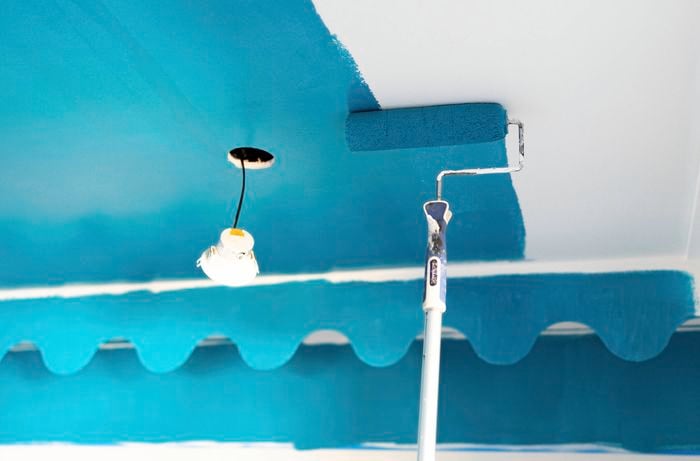 A person is applying vibrant blue paint with a paint roller along the upper wall and ceiling edge to create a colour-capping feature.