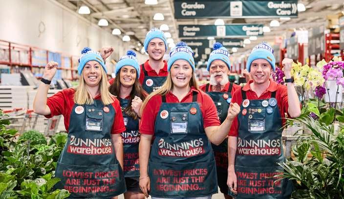 Seven store team members standing in a hardware and garden store aisle, wearing red shirts, green Bunnings Warehouse aprons and blue beanies, raising clenched fists. Indoor plants surround the group, with garden care signage visible overhead.