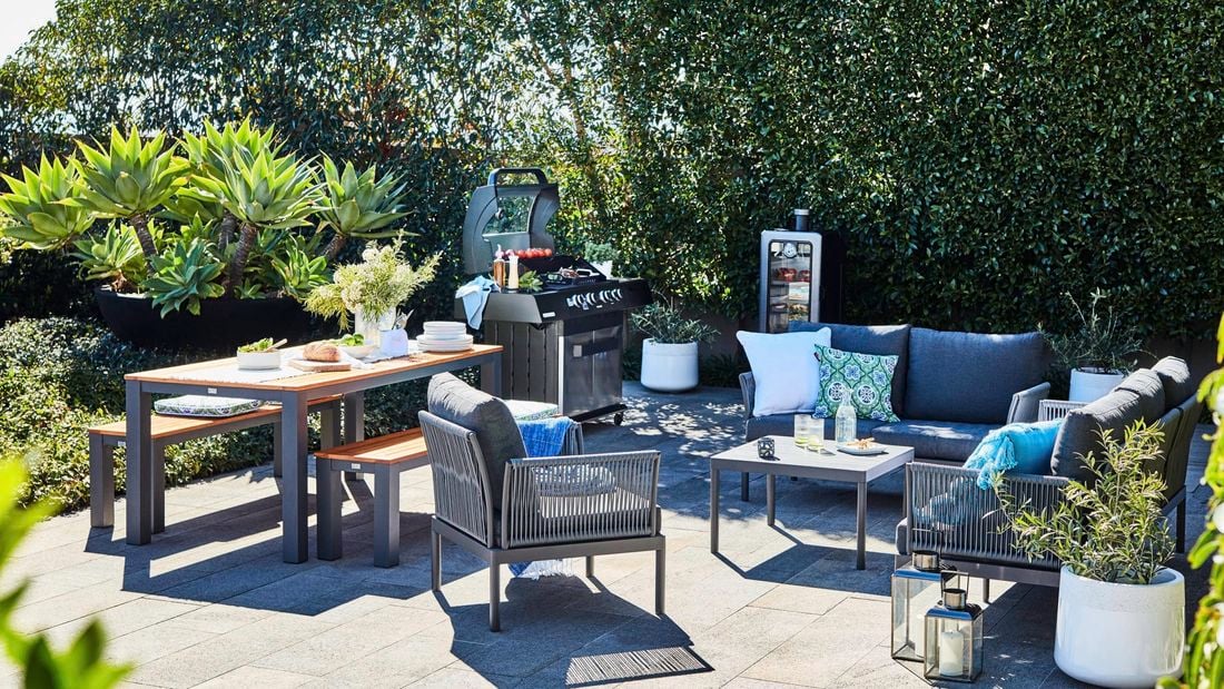 Outdoor entertaining area with a BBQ, plants, outdoor table and chairs in it.