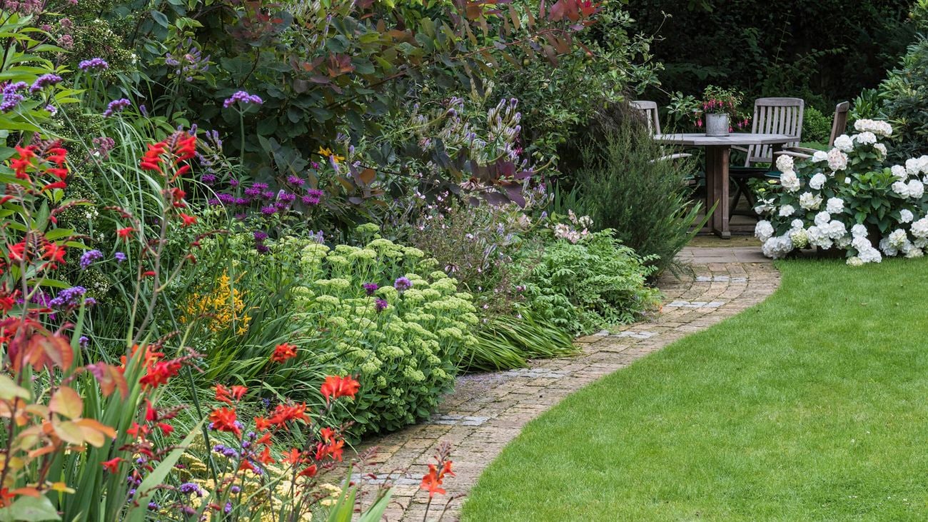  A colourful, blooming garden design with a paved footpath beside it, and a timber outdoor setting at the end of the path.