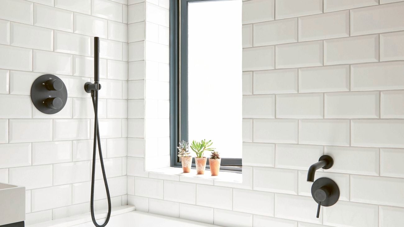 A bathroom with white subway tiles, a white bathtub, black shower fixtures, and a window above the tub.