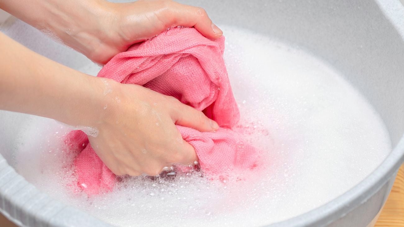 Hands washing pink clothing in bucket with soapy water.