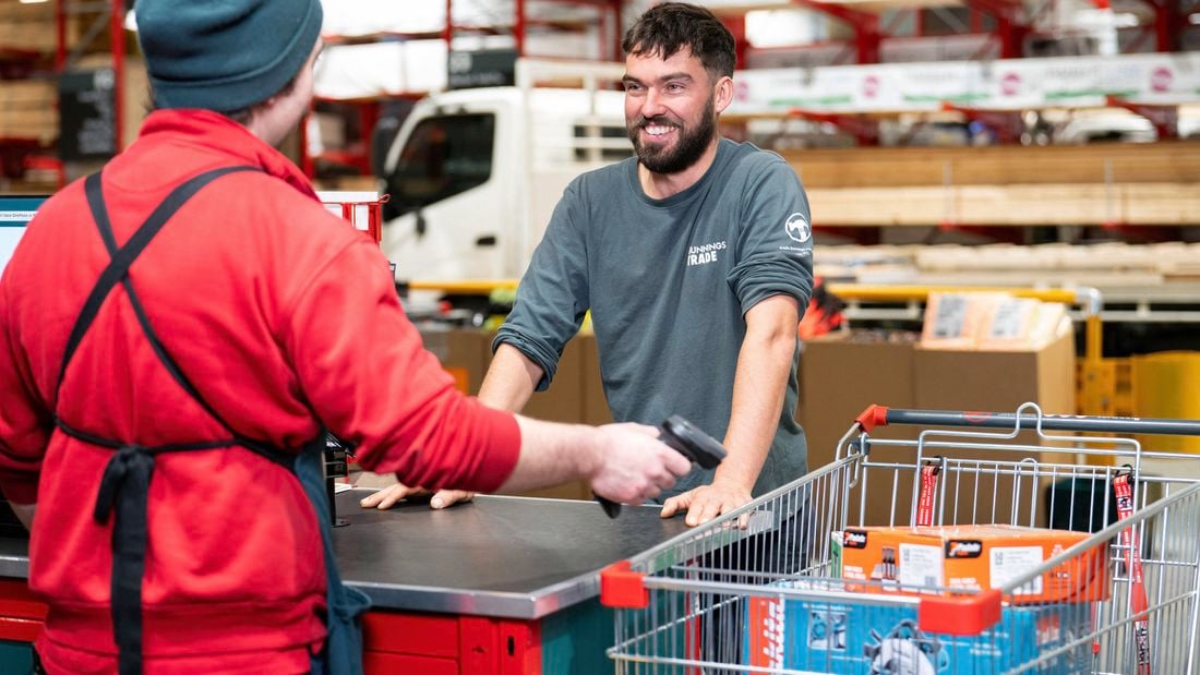 A man in a Bunnings Trade long-sleeved shirt smiles at a Bunnings Warehouse team member, who is scanning products in his trollery.