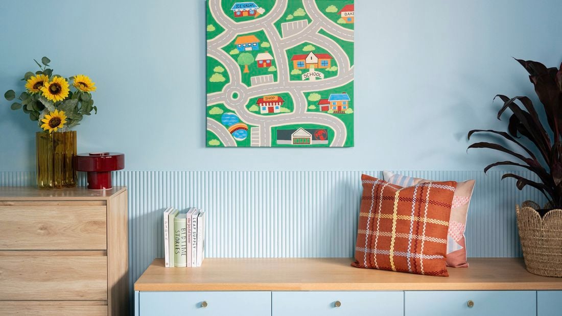 A light blue room with a map-themed wall art piece above a low cabinet decorated with books, pillows, plants, and sunflowers in a vase.