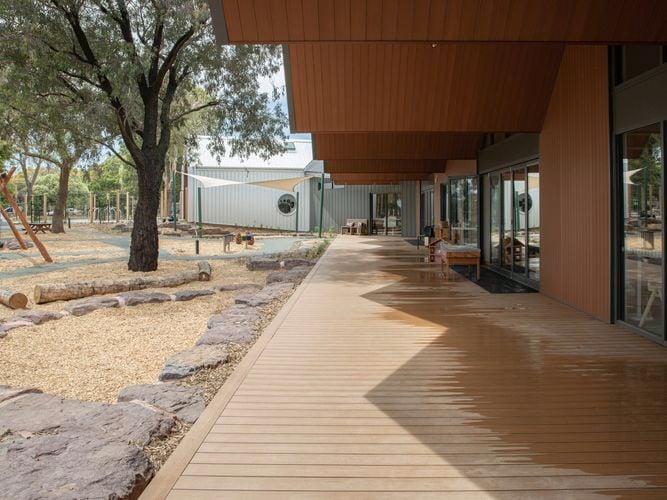 Outdoor wooden walkway with overhanging roof, landscaped garden on the left, and glass doors on the right leading into a modern building.