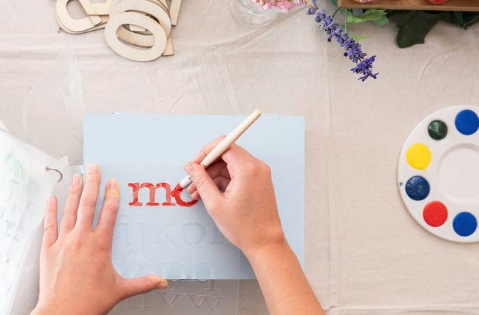 A person is using a stencil and paintbrush to apply colour and lettering while decorating a personalised DIY memory box.