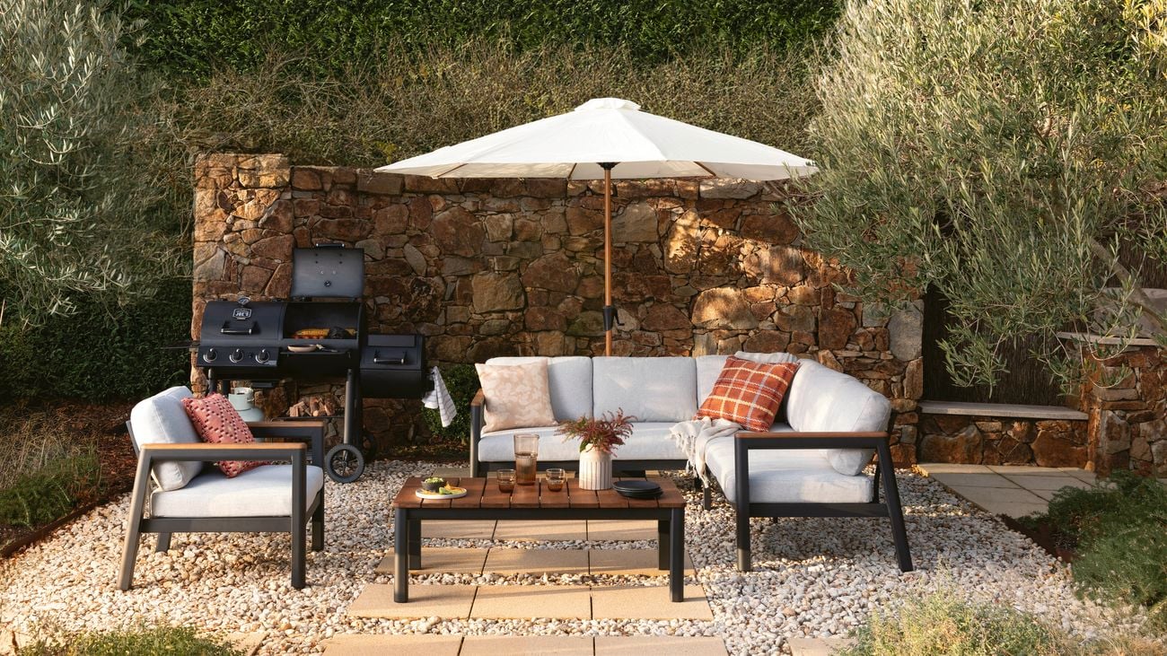   Outdoor seating area with a stone wall backdrop, featuring a light gray cushioned sofa and matching armchairs with black frames, a wooden coffee table with drinks and decor, a white patio umbrella, and a black barbecue grill on a gravel surface surrounded by greenery.