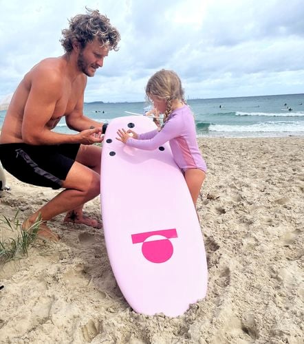 Father and young daughter fitting fins to a pink soft surfboard on the sand.