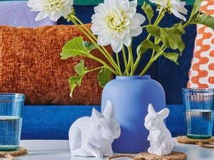 Two small bunnies made by plaster sitting in front of a blue vase with colourful cushions in the background