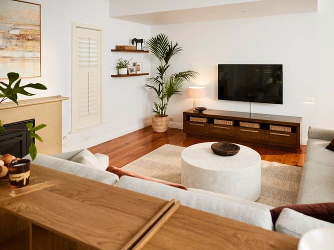 Light-filled living room with wooden floors, a wall-mounted TV above a low timber cabinet with baskets, a round coffee table on a rug, neutral sofas, indoor plants, and minimalist wall shelves.