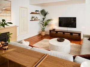 Light-filled living room with wooden floors, a wall-mounted TV above a low timber cabinet with baskets, a round coffee table on a rug, neutral sofas, indoor plants, and minimalist wall shelves.