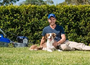A man with his dog, and a mower, sit on the backyard in the sun.
