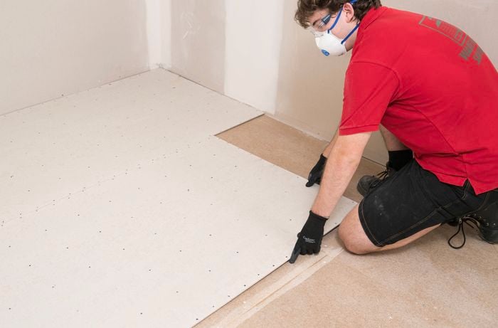 A Bunnings Team Member wearing safety gear is installing an underlay sheet in a brick pattern, running them opposite to the existing floorboards. 