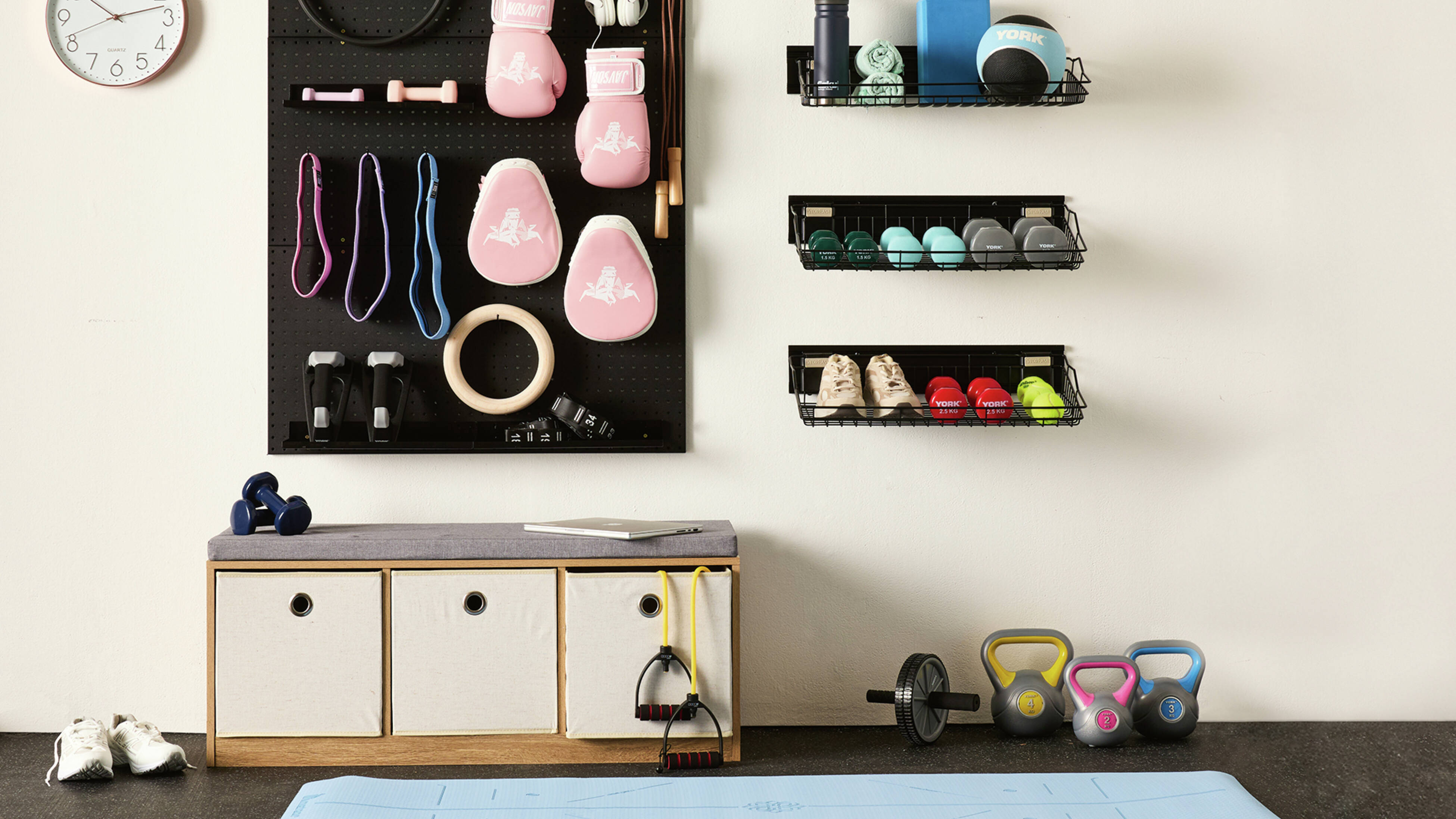 A white wall with three shelving baskets and a pegboard filled with home gym equipment including boxing gloves and weights. A yoga mat, kettle bells, sports shoes and bench seat are also in shot