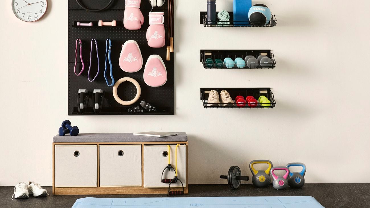 A white wall with three shelving baskets and a pegboard filled with home gym equipment including boxing gloves and weights. A yoga mat, kettle bells, sports shoes and bench seat are also in shot