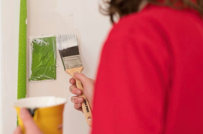 A person in a red T-shirt is using a paintbrush with precision, applying paint to the walls with the help of painter's tape.