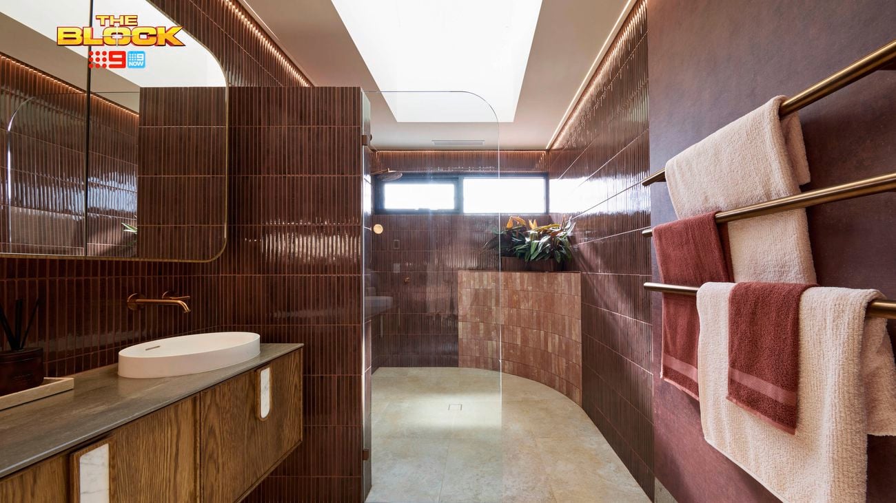 Modern bathroom with wooden vanity, oval sink, brass fixtures, glass shower, maroon tiles, towel racks, and skylight.