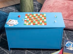 A finished DIY games storage box painted blue with a snakes and ladders board on the lid, styled with dice, playing cards and game pieces.