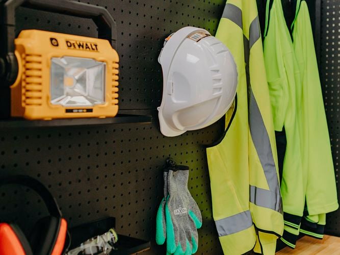 Black pegboard wall displaying safety gear including a yellow DeWalt work light, white hard hat, green and gray gloves, two high-visibility jackets, ear protection, and safety goggles, with a wooden floor beneath