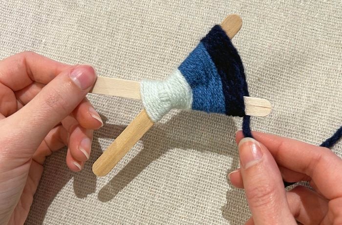 A left hand holds a paddle pop stick in place as the other one holds black yarn.