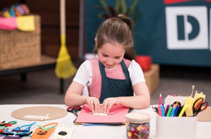A young girl is creating the base of her Halloween character by gluing popsicle sticks together