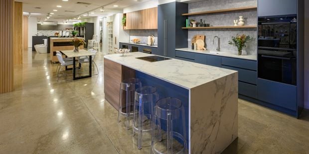 Contemporary kitchen showroom featuring a marble island with clear stools, navy cabinetry, built-in ovens, and a styled dining area.