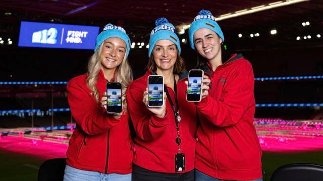 Three bunnings team members in red jackets and blue beanies stand in a brightly lit stadium, each holding up a smartphone displaying a promotional app or game. A large digital sign in the background shows “NR12” and “FightMND,” while the stadium seating is illuminated with pink lighting.