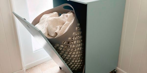 A light blue tilt-door laundry hamper cabinet with a grey hamper inside, holding white laundry.