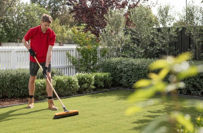 A Bunnings Team Member is using a stiff-bristled broom to brush the turf against the direction of the turf's grain