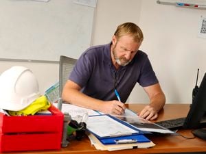 A man works at a home-office desk, with a construction hard hat sitting on his intray