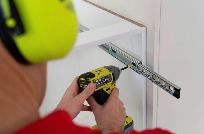 A person wearing ear protection is using a power drill to fix the outer runners inside the cabinet 