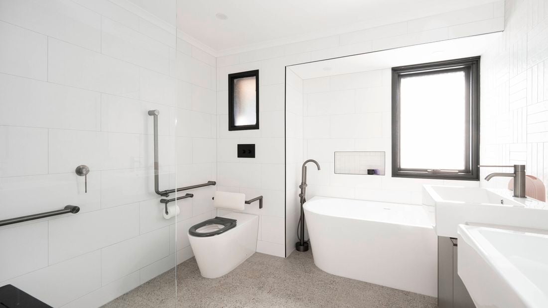 A white tiled bathroom with a bath under a framed window with handrails and toilet with handrails, a sink and a free standing shower