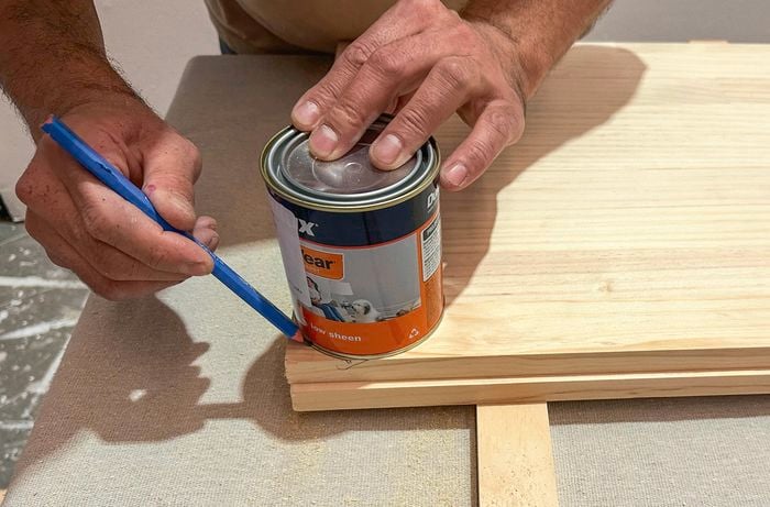 A hand uses a pencil to trace a curve around a paint tin sitting on a timber panel