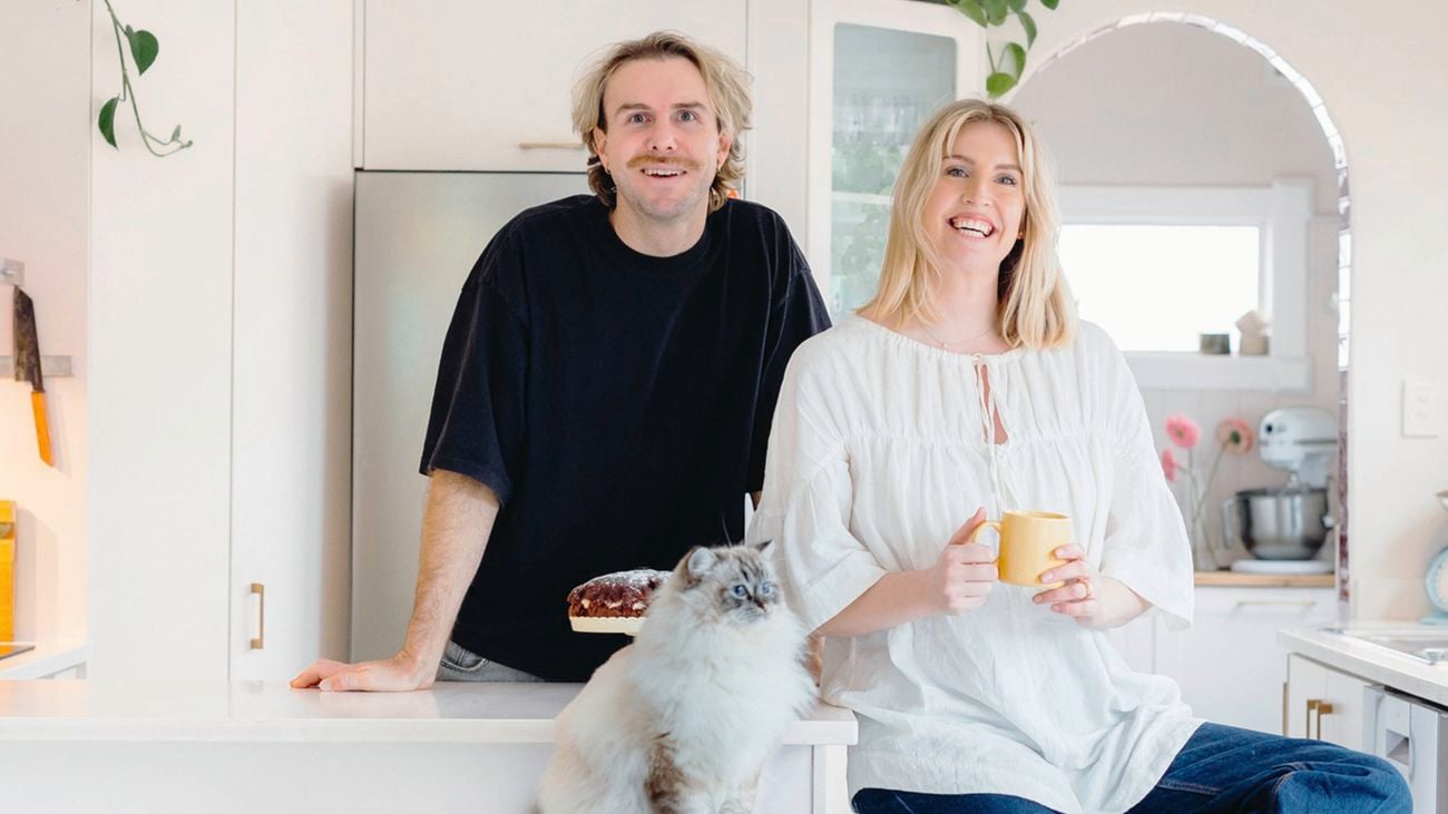 A man in a black t-shirt, a woman holding a yellow mug, and a fluffy cat sit in a bright kitchen.