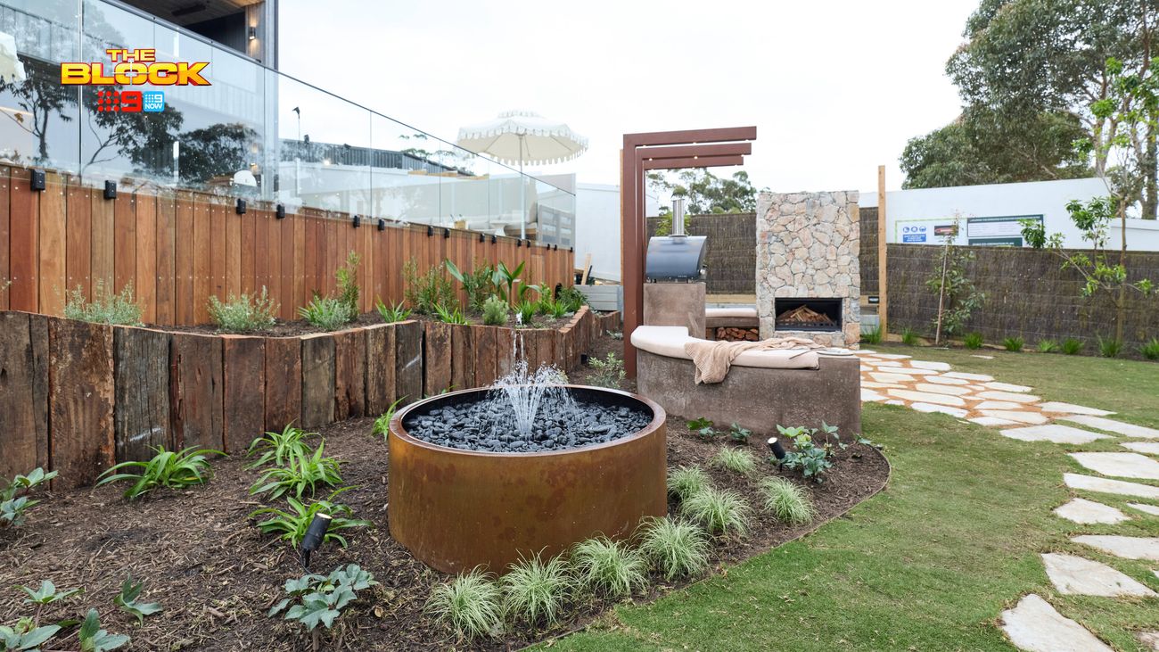 Landscaped outdoor garden featuring a circular rust-coloured water feature, raised timber garden beds, stone stepping paths and a built-in fireplace with seating.